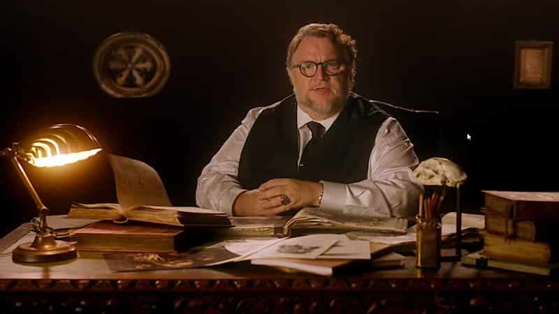 Guillermo del Toro speaks about his new Netflix horror anthology series, Cabinet of Curiosities-min Cabinet of Curiosities, Guillermo del Toro wearing a white shirt sitting behind at a desk full of books and papers and a night lamp