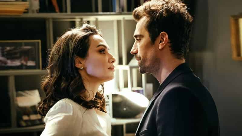 The first season of Kuş Uçuşu, which brought together Birce Akalay, İbrahim Çelikkol, and Miray Daner, attracted a large audience on Netflix - Season 2 it's coming! Here is what we know so far.-min Season 2 Kuş Uçuşu As the Crow Flies, Birce Akalay with dark brown hair wearing a white blouse standing in front and looking into the eyes of İbrahim Çelikkol who wears a blue suit